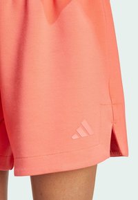 Krótki spodenki sportowe w kolorze brzoskwiniowym, wykonane z miękkiego materiału, z bocznym rozcięciem i delikatnym wytłoczonym logo Adidas na dolnym szwie.