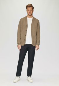 Beige gebreid vest met een geribde kraag, zes zwarte knopen, gedragen over een wit shirt. Gecombineerd met donkere broeken en witte sneakers.