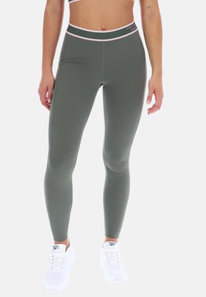 Femme portant des leggings taille haute vert olive avec le logo Ted Baker sur la ceinture et des baskets blanches Reebok, debout sur un fond blanc.