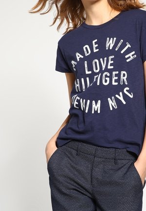 Kvinde iført navyblå T-shirt med hvid tekst "Made with Love Hilfiger Denim NYC" og mørke teksturerede bukser med hænderne i lommerne.