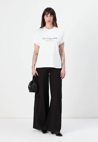 White oversized T-shirt with black logo print, paired with black wide-leg cargo pants featuring side pockets; model holds a black handbag.