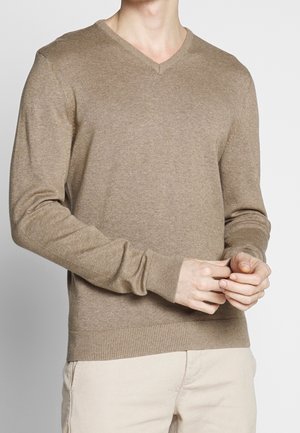 Pull à col en V, à manches longues, en maille brun clair. Il présente des poignets et un ourlet côtelés. Le modèle porte un pantalon de couleur crème.