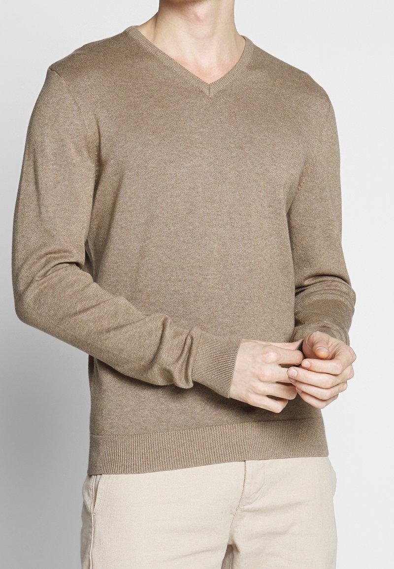 Pull à col en V, à manches longues, en maille brun clair. Il présente des poignets et un ourlet côtelés. Le modèle porte un pantalon de couleur crème.