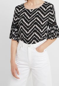 Schwarzes Oberteil mit weißem floralem Zickzackmuster, runder Halsauschnitt und ausgestellten Ärmeln. Kombiniert mit hochtaillierten weißen Jeans.