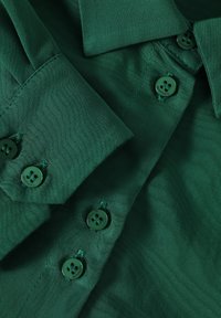 Gros plan sur un tissu vert foncé avec des boutons, montrant le col et le poignet de la chemise avec des détails de boutons et des coutures.