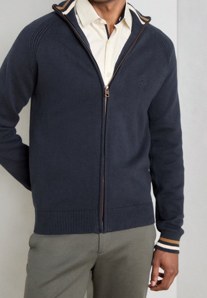 Homme portant un pull zippé bleu foncé sur une chemise crème et un pantalon olive, debout avec une main le long du corps sur un fond clair.