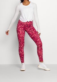 Leggings a stampa leopardo rosa in un materiale elasticizzato, abbinati a una maglietta bianca ampia. Modella che indossa scarpe bianche leggere.