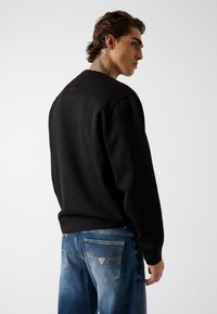 Czarna bluza z gładką fakturą, zestawiona z jasnoniebieskimi dżinsami z naszywką z logo na tylnej kieszeni.