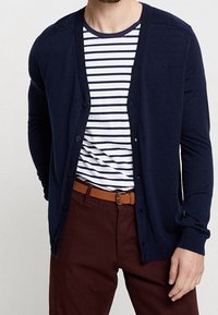 Man som bär en marinblå knäppbar kofta över en vit och marinrandig t-shirt, brun bälte och vinröda byxor, stående med en hand synlig.