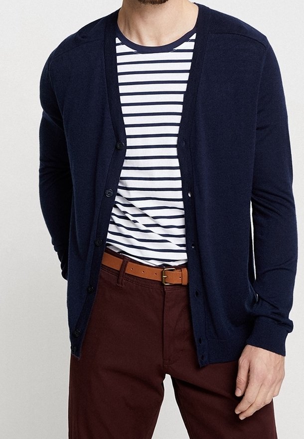 Man som bär en marinblå knäppbar kofta över en vit och marinrandig t-shirt, brun bälte och vinröda byxor, stående med en hand synlig.