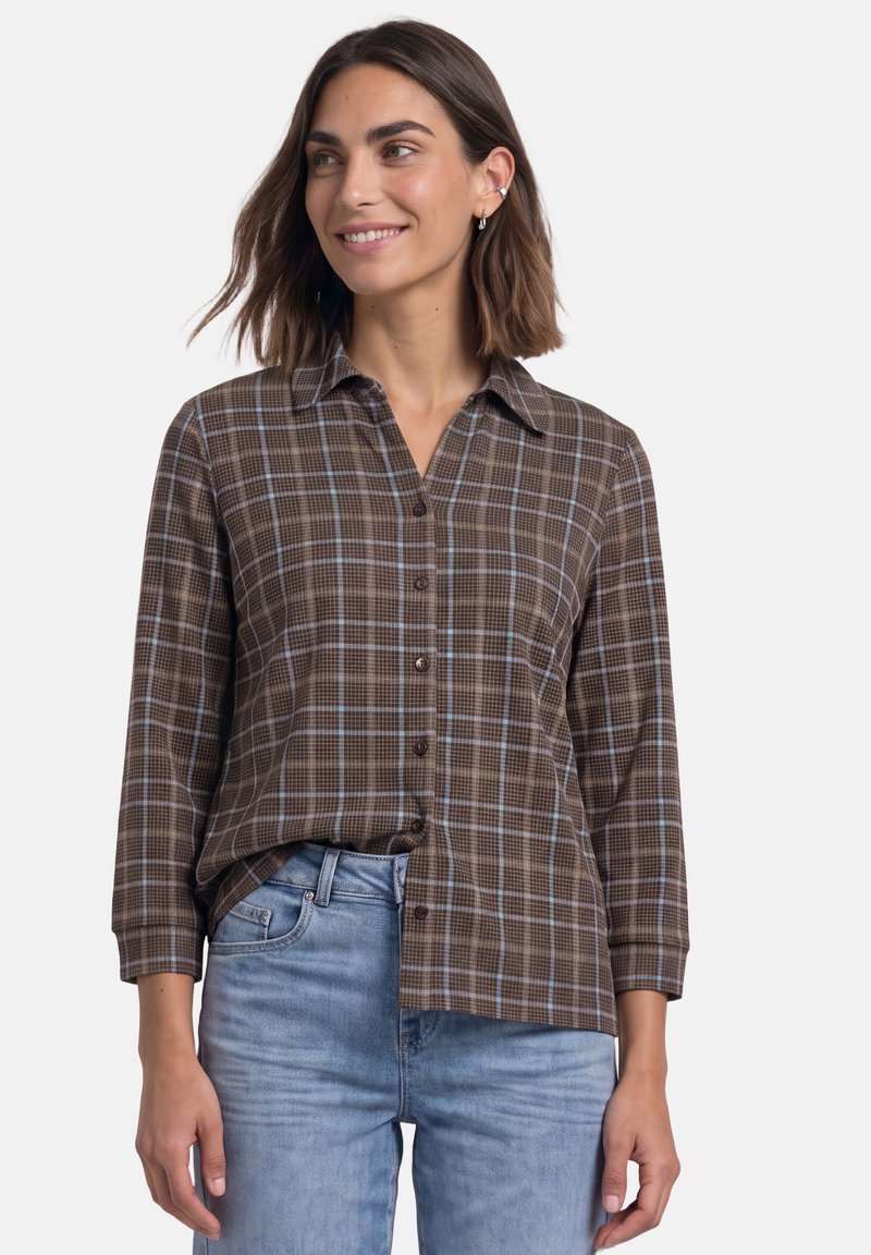 Femme portant une chemise à carreaux marron boutonnée avec des manches trois-quarts et un jean bleu clair, souriante et regardant sur le côté.