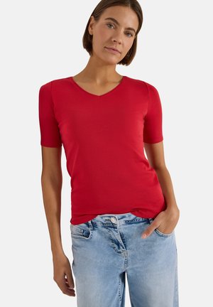 Femme aux cheveux bruns courts portant un t-shirt rouge à manches courtes et col en V ainsi qu'un jean bleu clair, une main dans la poche, sur un fond uni.