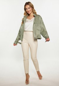 Jachetă bomber verde măsliniu cu mâneci lungi, top din satin crem, colanți din piele ecologică bej și pantofi cu toc aurii cu curele. Croi lejer, închidere cu fermoar.
