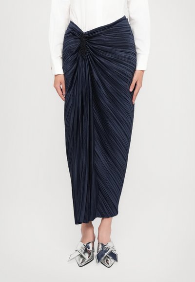 Gonna pieghettata blu navy con dettaglio torcigliato in vita. Presenta una texture liscia e lunghezza alla caviglia, abbinata a scarpe metallizzate argentate.