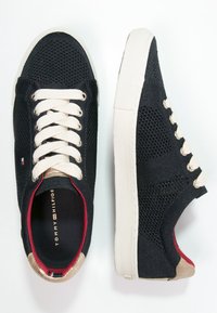 Sneaker in rete nera con lacci crema, dettagli dorati e rivestimento interno rosso. Presenta una suola testurizzata e un design a taglio basso.
