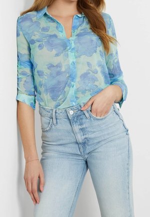 Vrouw draagt een doorschijnende blauwe bloemenblouse met knopen en opgerolde mouwen, en lichtblauwe jeans met hoge taille, hand in de voorzak.