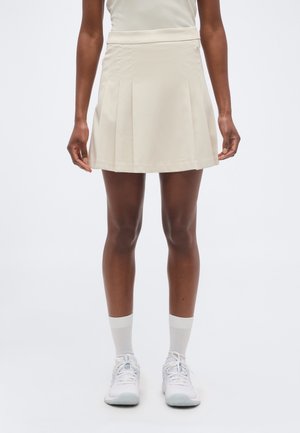 Mujer con falda beige plisada, calcetines blancos y zapatillas blancas, de pie frente a un fondo liso y claro.