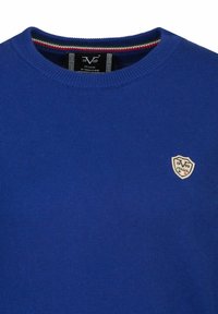 Blauer Strickpullover mit Rundhalsausschnitt, geripptem Kragen und kleinem Wappenlogo auf der Brust, das von roten und grünen Akzentstreifen am Kragen umgeben ist.