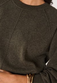 Maglione verde oliva a maglia con collo a girocollo a coste, maniche raglan e una texture sottile. Bracciali dorati visibili al polso.