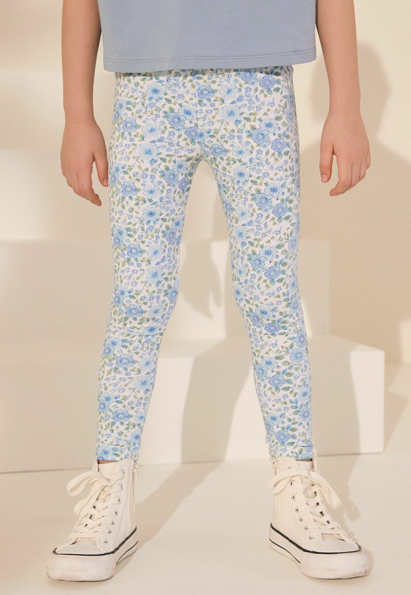 Niño con leggings blancos con estampado floral azul y zapatillas blancas altas, de pie contra un fondo claro.