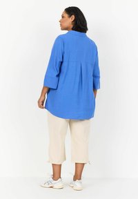 Camisa de lino azul con mangas tres cuartos, detalle de pliegue en la espalda, combinada con pantalones cortos beige y zapatillas blancas.