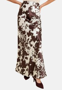 Jupe maxi en satin présentant un imprimé vache marron et blanc, texture lisse et taille élastique. Portée avec des talons bordeaux.
