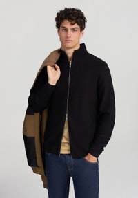 Giovane uomo che indossa un maglione nero con zip e jeans blu, che tiene una giacca marrone sulla spalla, davanti a uno sfondo neutro.