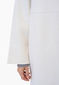 Weißer Wollmantel mit strukturiertem Finish, ausgestattet mit langen Ärmeln und einem grauen Strickbündchen, das darunter hervorschaut. Schlichtes Design mit einer seitlichen Tasche.
