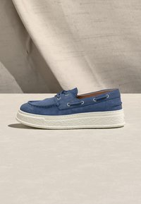 Μπλε σουέντ μοκασίνι με λευκή, υφή, χοντρή σόλα, λεπτομέρειες με κορδόνια και καφέ εσωτερική επένδυση, τοποθετημένο σε φωτεινή επιφάνεια με φόντο μπεζ ύφασμα.