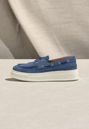 Blaues Wildleder-Bootsschuh mit weißer, strukturierter, dicker Sohle, Schnürdetails und beigem Innenfutter, auf heller Oberfläche vor beigem Stoffhintergrund platziert.