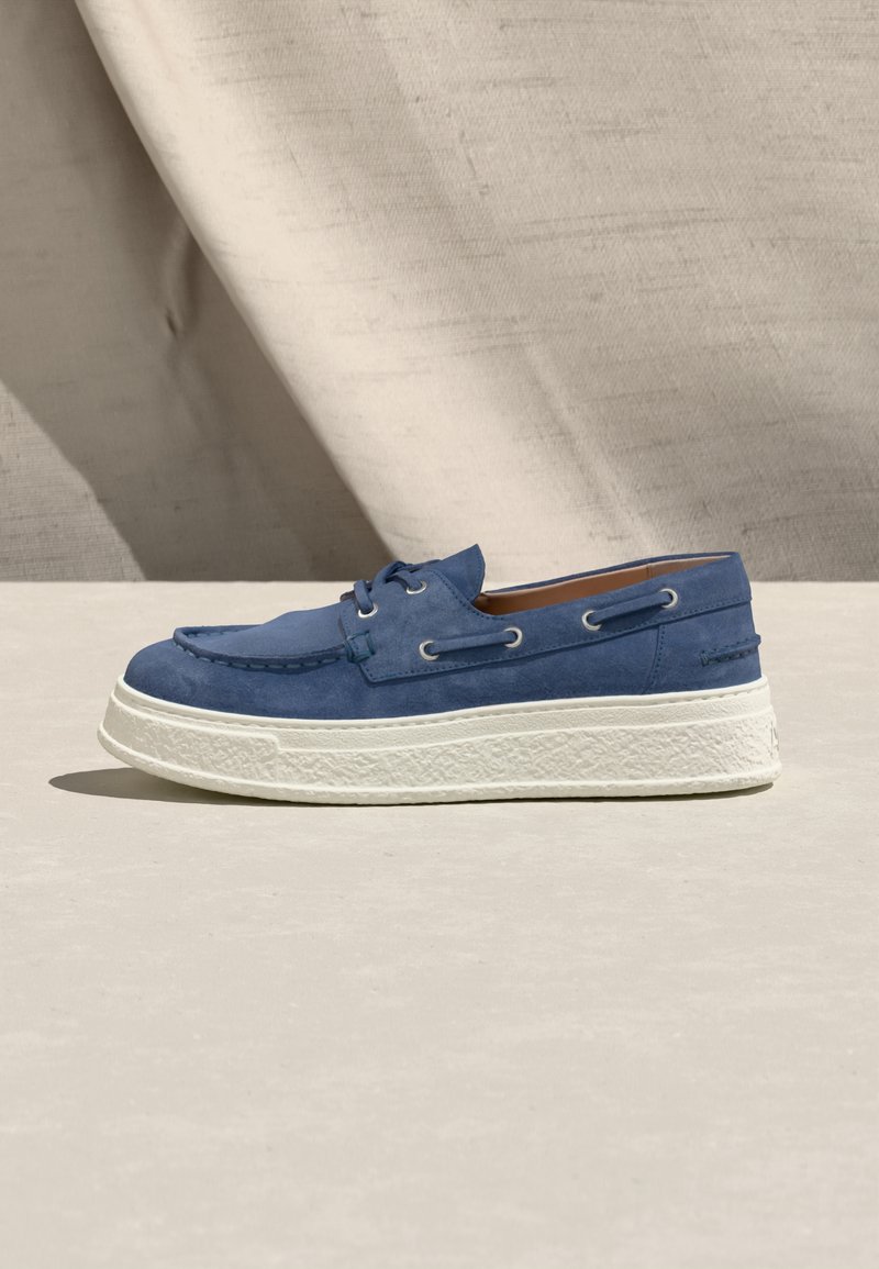 Μπλε σουέντ μοκασίνι με λευκή, υφή, χοντρή σόλα, λεπτομέρειες με κορδόνια και καφέ εσωτερική επένδυση, τοποθετημένο σε φωτεινή επιφάνεια με φόντο μπεζ ύφασμα.