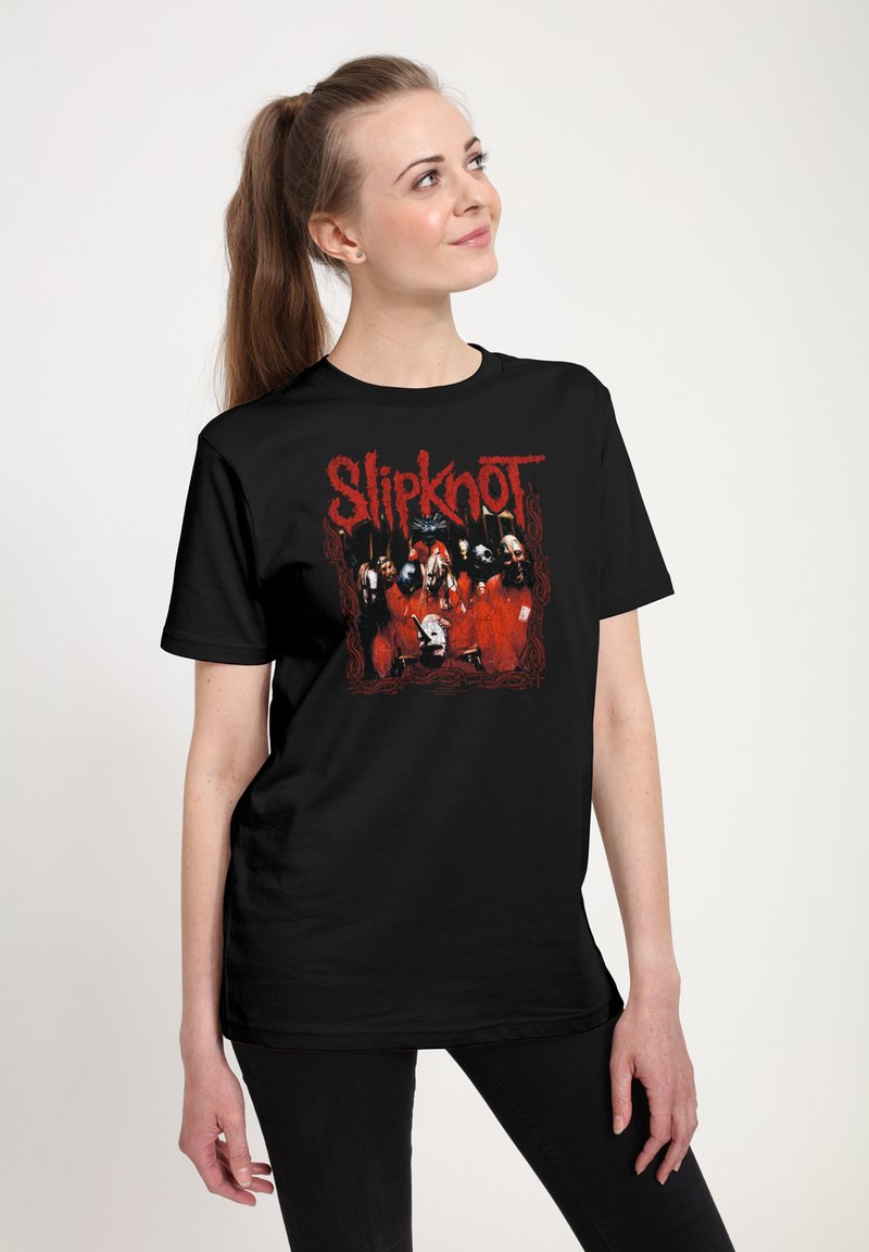 Young woman with a ponytail wearing a black Slipknot band T-shirt featuring masked members in red attire on a light background.