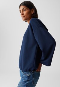 Donna con capelli corti e scuri, che indossa un top a maniche lunghe blu navy con texture e jeans blu, in piedi di profilo su uno sfondo chiaro e uniforme.