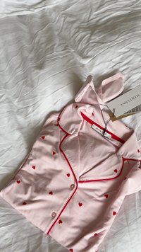 Lyserød pyjamasbluse med røde hjerteprints og røde kantbånd, foldet på hvidt krøllet lagen, med "Anna Field" mærketiketter fastgjort.