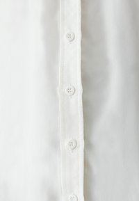 Camicia bianca con bottoni, caratterizzata da una consistenza liscia e una patta con sei bottoni abbinate sul davanti. Design semplice e classico.