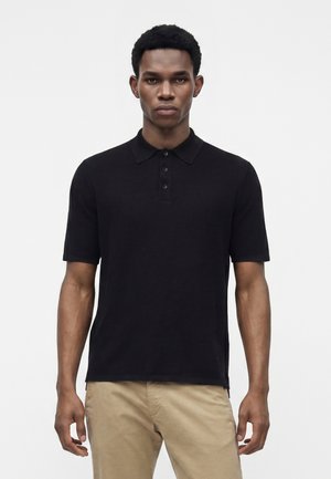 Mand iført en sort kortærmet poloshirt og beige bukser står mod en ensfarvet hvid baggrund.