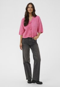 Rosa gestrickte Strickjacke mit kurzen Ärmeln und drei blumenförmigen Knöpfen. Kombiniert mit grauen hoch taillierten Jeans und schwarzem Schuhwerk.