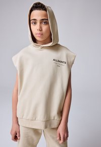 Felpa beige senza maniche con cappuccio e orlo a coste, presenta una stampa del logo sul petto. Indossata con pantaloni beige abbinati, di tessuto liscio.