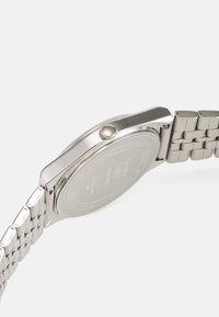 Silverfärgad klocka i rostfritt stål med rund klockkapsel, polerad yta och strukturerat metallarmband. Sidokrona och graverad baksida synlig.
