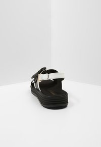 Mustat ja valkoiset platform-sandaalit, joissa on useita hihnoja ja kultainen solkiyksityiskohta. Jalkapohja on tumma, teksturoitu ja pehmustettu.