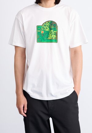 Personne portant un t-shirt blanc avec un motif abstrait de circuit imprimé vert et orange centré sur la poitrine, associé à un pantalon noir.