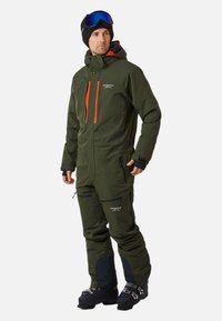 Zielony wodoodporny kombinezon narciarski z pomarańczowymi akcentami, wieloma zamkami błyskawicznymi, regulowanymi mankietami i wzmocnionymi łatami. Noszony z butami narciarskimi i goglami.