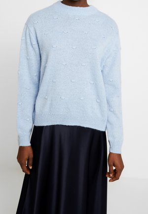 Light blue, textured knit sweater with small bobble details worn with a black satin skirt, shown from mid-chest to hips.