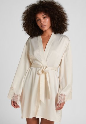 Frau mit lockigem Haar, die einen cremefarbenen Seidenmorgenmantel mit Spitzenmanschetten trägt, der in der Taille gebunden ist, steht vor einem schlichten Hintergrund.