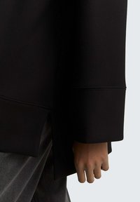 Hand van een persoon in een zwart, lang mouwend kledingstuk met een uitgebreide manchette, rustend nabij grijze broek, tegen een effen lichte achtergrond.