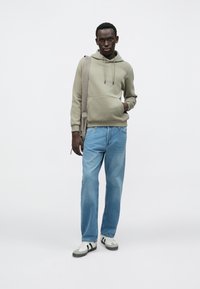 Lys grønn hettegenser med kengurulomme, lyseblå denimjeans og grå/hvite joggesko med svarte striper. Modell holder en bag.