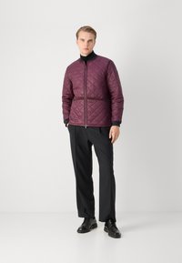 Quilted burgundy jacket with a front zipper and black accents, paired with black trousers and black shoes, set against a neutral background.