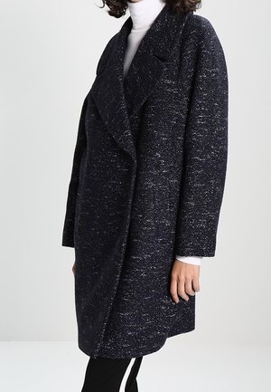 Femme portant un long manteau texturé foncé sur un col roulé blanc, debout avec la main gauche détendue le long du corps.