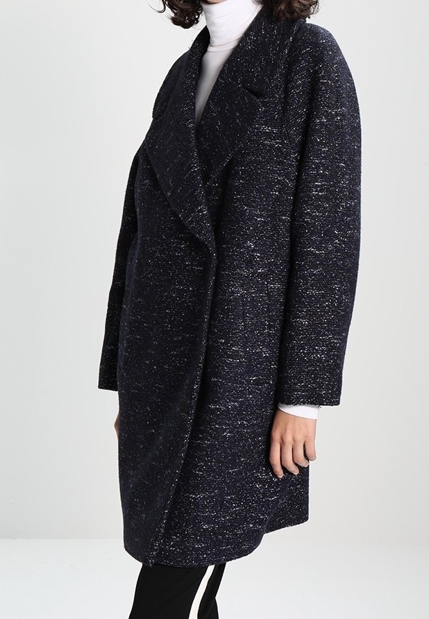 Femme portant un long manteau texturé foncé sur un col roulé blanc, debout avec la main gauche détendue le long du corps.