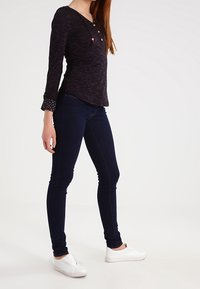 Frau in einem dunklen Langarmoberteil, dunklen Skinny-Jeans und weißen Slippern, die vor einem schlichten hellen Hintergrund steht.